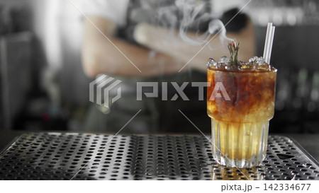 Smoking craft cocktail featuring a rosemary garnish, ice, and layers of liquor, served on a bar mat with a bartender preparing drinks Smoking craft cocktail featuring a rosemary garnish, ice, and layers of liquor, served on a bar mat with a bartender preparing drinks 142334677