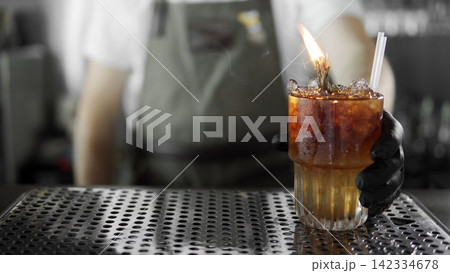 Bartender serving flaming craft cocktail with ice and garnish in a crystal glass on a bar counter Bartender serving flaming craft cocktail with ice and garnish in a crystal glass on a bar counter 142334678