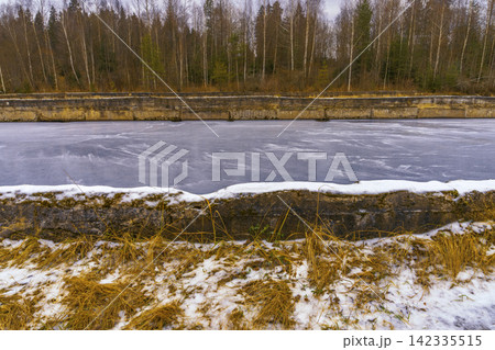Frozen canal with concrete embankments and leafless birch trees in late winter landscape Frozen canal with concrete embankments and leafless birch trees in late winter landscape 142335515