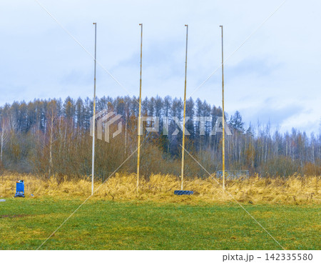 Four empty flagpoles standing on grassy field with autumn trees and pale overcast sky Four empty flagpoles standing on grassy field with autumn trees and pale overcast sky 142335580