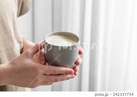 Human hands holding a cup of cappuccino coffee on a window background with morning shadow Human hands holding a cup of cappuccino coffee on a window background with morning shadow 142337734