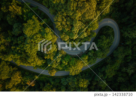 Aerial view of a winding road through a lush green forest with a driving car. Aerial view of a winding road through a lush green forest with a driving car. 142338710