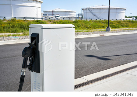 Modern electric vehicle charging station with industrial storage tanks in background Modern electric vehicle charging station with industrial storage tanks in background 142341348
