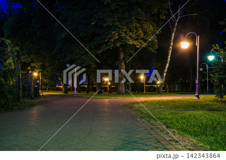 The tiled road in the night green park with lanterns in summer. Benches in the park during the summer season at night. Illumination of a park road with lanterns. Park of Eternal Glory. Ukraine The tiled road in the night green park with lanterns in summer. Benches in the park during the summer season at night. Illumination of a park road with lanterns. Park of Eternal Glory. Ukraine 142343864