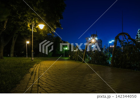 The tiled road in the night green park with lanterns in summer. Benches in the park during the summer season at night. Illumination of a park road with lanterns. Park of Eternal Glory. Ukraine The tiled road in the night green park with lanterns in summer. Benches in the park during the summer season at night. Illumination of a park road with lanterns. Park of Eternal Glory. Ukraine 142344050