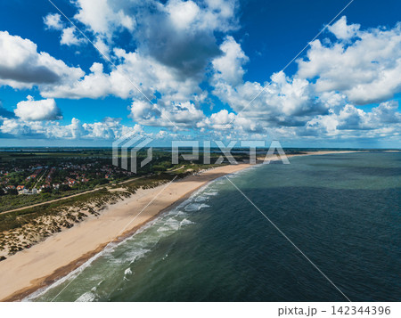 High angle wide horizontal aerial drone photograph of North Sea coastline, sandy beach, sand dunes, coastal resort village, and cloudy sky at Haamstede, Zeeland, Netherlands on sunny summer day 142344396