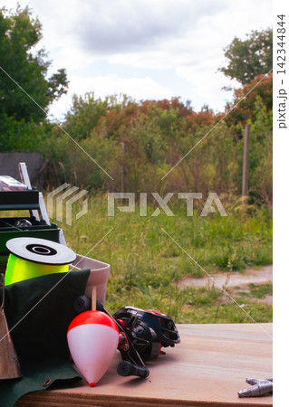 Fishing equipment on a wooden surface with a bobber, reel, and spool of line, preparing for an outdoor angling hobby in a natural setting Fishing equipment on a wooden surface with a bobber, reel, and spool of line, preparing for an outdoor angling hobby in a natural setting 142344844
