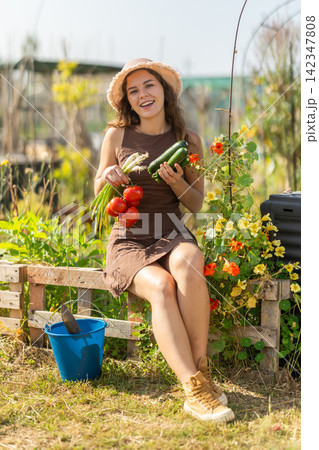 Positive woman in dress posing with tomatoes, zucchini and green onions in garden 142347808