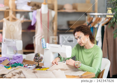 Working in sewing workshop - female dressmaker working at sewing machine making stitches on fabric Working in sewing workshop - female dressmaker working at sewing machine making stitches on fabric 142347826