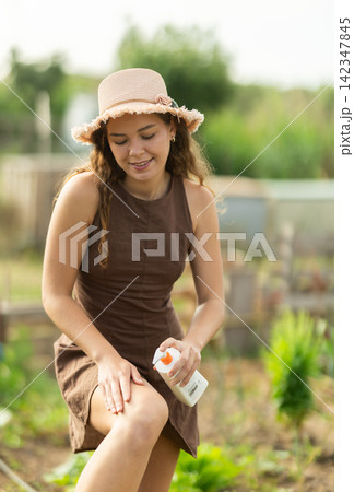 Smiling girl in dress and hat applying sunscreen to skin while gardening Smiling girl in dress and hat applying sunscreen to skin while gardening 142347845