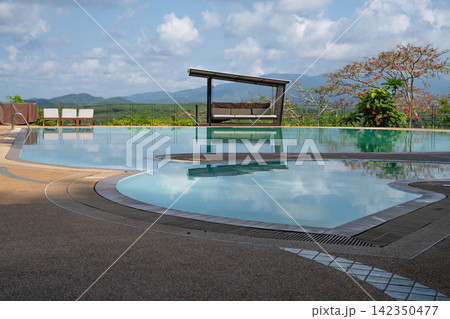 Outdoor swimming pool overlooking green mountains and tropical countryside beneath a bright cloudy sky. Scenic vacation setting for relaxation, wellness and resort travel. Relaxing outdoor retreat. Outdoor swimming pool overlooking green mountains and tropical countryside beneath a bright cloudy sky. Scenic vacation setting for relaxation, wellness and resort travel. Relaxing outdoor retreat. 142350477