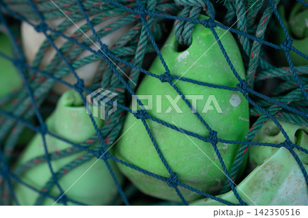 Closeup of green fishing floats and rope secured beneath a blue net on commercial fishing equipment. Durable marine gear used by fishers at sea for net buoyancy, handling and seafood operations. 142350516