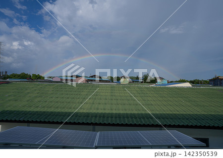 Full rainbow arching across sky above rooftops and photovoltaic solar panels, combining natural weather beauty with renewable energy and sustainable urban living. Renewable energy infrastructure. Full rainbow arching across sky above rooftops and photovoltaic solar panels, combining natural weather beauty with renewable energy and sustainable urban living. Renewable energy infrastructure. 142350539