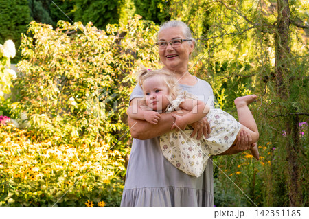 Smiling Grandmother Carrying Granddaughter in Sunny Garden Smiling Grandmother Carrying Granddaughter in Sunny Garden 142351185