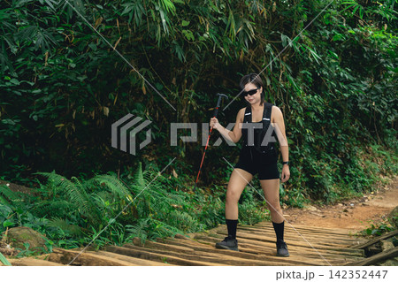 Woman with black outfit and sunglasses hiking on wooden path surrounded by lush green forest vegetation in daytime Woman with black outfit and sunglasses hiking on wooden path surrounded by lush green forest vegetation in daytime 142352447