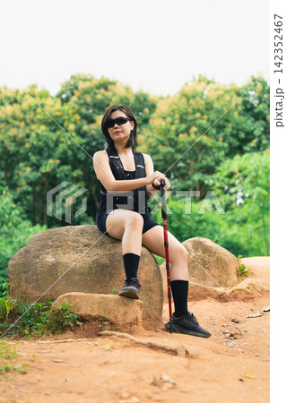 Young woman in casual black outfit hiking and resting on large stone with walking stick in green forest park Young woman in casual black outfit hiking and resting on large stone with walking stick in green forest park 142352467
