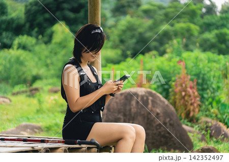 Young woman in black outfit sitting outdoors on wooden bench looking at smartphone beneath rustic shelter in nature Young woman in black outfit sitting outdoors on wooden bench looking at smartphone beneath rustic shelter in nature 142352497