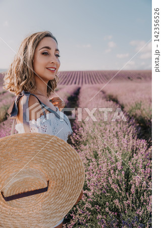 Woman lavender field smiling while holding a large straw hat in a blooming purple flower garden under blue sky Woman lavender field smiling while holding a large straw hat in a blooming purple flower garden under blue sky 142356526