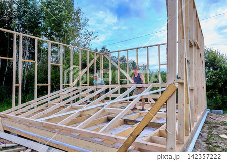 Wall raising shows two builders manually lifting completed wall frame into place as timber frame house takes shape during construction. Wall raising shows two builders manually lifting completed wall frame into place as timber frame house takes shape during construction. 142357222
