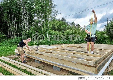 Foundation on concrete blocks supports timber frame construction, workers assemble wooden floor structure during building process. Foundation on concrete blocks supports timber frame construction, workers assemble wooden floor structure during building process. 142357224