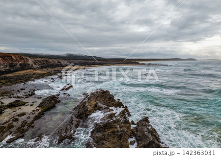 Aerial view of ocean waves crashing onto the rugged rocky coastline at Lake Arragan Beach beneath a cloudy sky, showcasing dramatic surf, exposed rock formations and the wild Pacific coastline  142363031