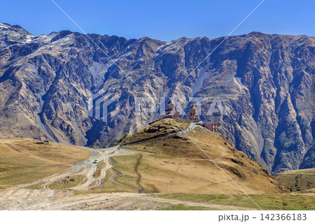 Gergeti Trinity Church stands against Caucasus mountains in Georgia. Scenic distant back view under bright morning sky 142366183