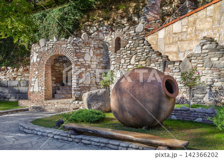 Big clay qvevri vessels lie in a green garden by a stone wall, Georgia. Entrance to a traditional winery in daylight Big clay qvevri vessels lie in a green garden by a stone wall, Georgia. Entrance to a traditional winery in daylight 142366202