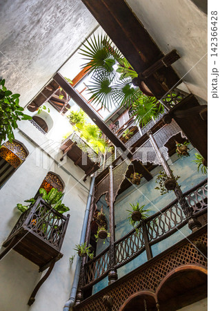 Looking up at the open-air central courtyard of a traditional Arabic riad featuring ornate carved wooden balconies, lush potted plants, and classic architectural arches. 142366428
