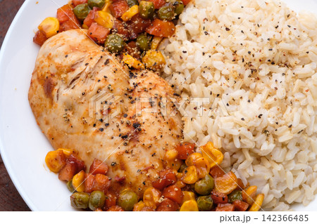a dish of homemade roasted chicken breast and dice carrots, beans, corns with brown rice on dark red wooden table and cutlery a dish of homemade roasted chicken breast and dice carrots, beans, corns with brown rice on dark red wooden table and cutlery 142366485