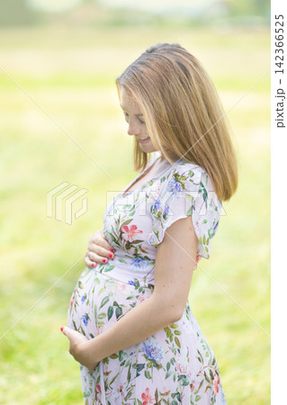 Side profile portrait of a smiling pregnant woman in a floral print summer dress holding her baby bump and looking down lovingly in an outdoor park setting Side profile portrait of a smiling pregnant woman in a floral print summer dress holding her baby bump and looking down lovingly in an outdoor park setting 142366525