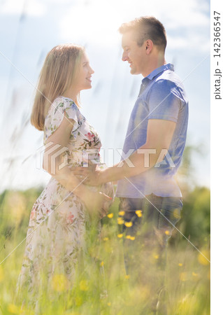 Side view of a happy pregnant couple embracing gently and holding the belly in a sunlit grassy field during summer. Romantic maternity concept of love, family preparation, and new life. Side view of a happy pregnant couple embracing gently and holding the belly in a sunlit grassy field during summer. Romantic maternity concept of love, family preparation, and new life. 142366547
