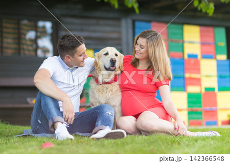 Happy young pregnant couple sitting on the grass in a park, smiling lovingly at their Golden Retriever dog positioned between them in front of a colorful backdrop Happy young pregnant couple sitting on the grass in a park, smiling lovingly at their Golden Retriever dog positioned between them in front of a colorful backdrop 142366548