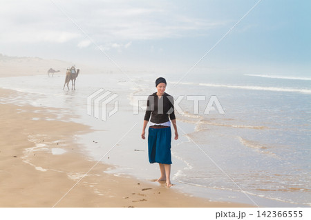 Woman wearing a black headscarf and long skirt walking barefoot along the wet shoreline of a foggy beach, with camels visible in the hazy distance Woman wearing a black headscarf and long skirt walking barefoot along the wet shoreline of a foggy beach, with camels visible in the hazy distance 142366555