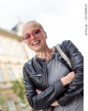 Smiling woman with pPortrait of a happy, stylish young woman in a black leather jacket and tinted red sunglasses, laughing with her arms crossed outdoors on a bright day 142366624