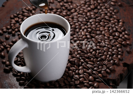 coffee droplets drop on black coffee water in white mug with surrounded by roasted coffee beans on dark red wood table 142366652