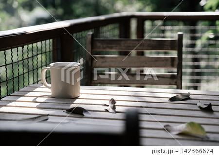 a coffee cup and many dried leaves on wooden table and chair at balcony with blurred green trees in background in afternoon a coffee cup and many dried leaves on wooden table and chair at balcony with blurred green trees in background in afternoon 142366674