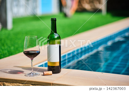 A bottle of red wine and a glass of wine are set on the edge of a clear blue pool, surrounded by a lush green lawn. The afternoon sun shines down, creating a relaxing and inviting atmosphere. A bottle of red wine and a glass of wine are set on the edge of a clear blue pool, surrounded by a lush green lawn. The afternoon sun shines down, creating a relaxing and inviting atmosphere. 142367003