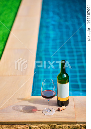 A bottle of red wine and a glass of wine are set on the edge of a clear blue pool, surrounded by a lush green lawn. The afternoon sun shines down, creating a relaxing and inviting atmosphere. 142367004