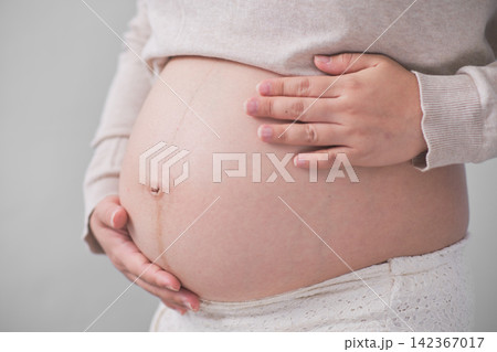 The mature Asian woman stood on the white backdrop, gracefully unveiling her large round belly, cradling it with both of her hands. The mature Asian woman stood on the white backdrop, gracefully unveiling her large round belly, cradling it with both of her hands. 142367017