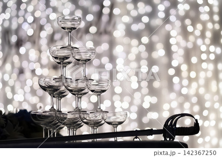 a silhouette shot of small tower of champagne's glass in wedding reception party near by a big cake and golden light bulbs in background. a silhouette shot of small tower of champagne's glass in wedding reception party near by a big cake and golden light bulbs in background. 142367250