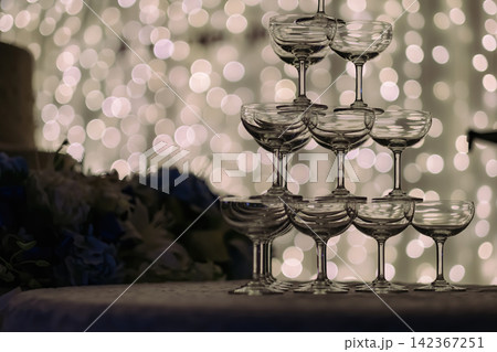 a silhouette shot of small tower of champagne's glass in wedding reception party near by a big cake and golden light bulbs in background. a silhouette shot of small tower of champagne's glass in wedding reception party near by a big cake and golden light bulbs in background. 142367251