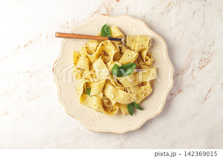 Fresh papardelle pasta, with basil and pesto sauce, is served, on a white plate, with a fork held by hand 142369015