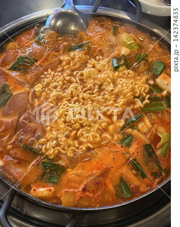 A hearty bowl of Budaejjigae (Korean Army Stew), filled with sausage, tofu, kimchi, and noodles in a spicy broth. This flavorful dish brings warmth and comfort, perfect for sharing. A hearty bowl of Budaejjigae (Korean Army Stew), filled with sausage, tofu, kimchi, and noodles in a spicy broth. This flavorful dish brings warmth and comfort, perfect for sharing. 142374835