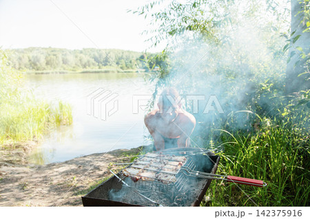 A young teenage boy stands behind a grill with his silhouette partially hidden by thick white smoke rising from juicy meat cooking on a wire rack. 142375916