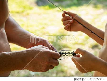 Close up of adult and child hands assembling a spring fishing feeder on a line outdoors during a summer day. Close up of adult and child hands assembling a spring fishing feeder on a line outdoors during a summer day. 142375930