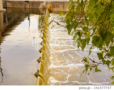 Water flows over a dam near a city on a sunny day 142377737
