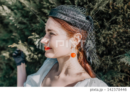 Redhead woman veil wearing a vintage hat and cherry earrings in a sunny garden setting while enjoying the golden hour 142378446