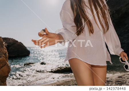 Woman beach fashion Young woman wearing white linen shirt holds a stone on a sunny coast with ocean waves Woman beach fashion Young woman wearing white linen shirt holds a stone on a sunny coast with ocean waves 142378448