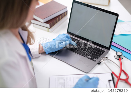 doctor typing on laptop at office desk with stethoscope and medical chart in blue and white setting over-the-shoulder view professional healthcare scene clean background. 142379259