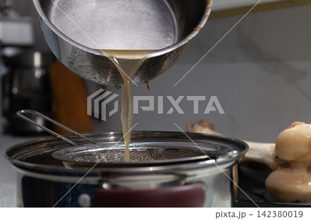 Pouring liquid from metal pot through mesh strainer into receiving pot in kitchen Pouring liquid from metal pot through mesh strainer into receiving pot in kitchen 142380019